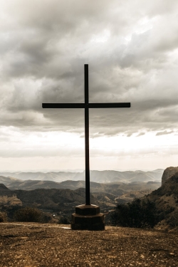 A powerful silhouette of a cross set against a dramatic cloudy mountain landscape.